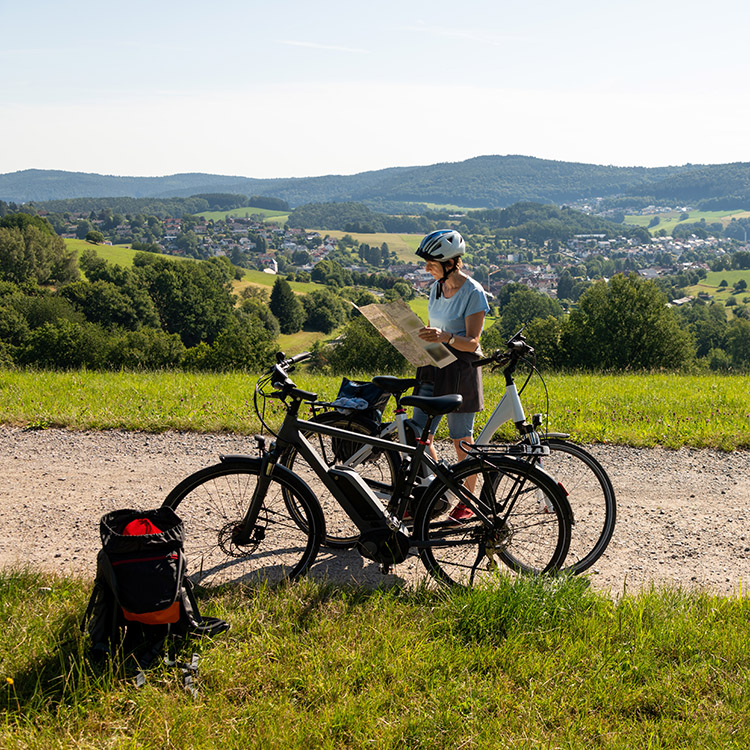 Frau mit Helm studiert Karte neben zwei modernen E-Bikes mit Panoramablick auf hügelige Landschaft – Diermann - Bergsieger GmbH